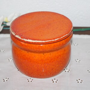 May include: A round, orange ceramic jar with a lid. The jar has a glossy finish with a textured surface. The lid has a white rim. The jar is sitting on a white, floral-patterned cloth. Decorative item.