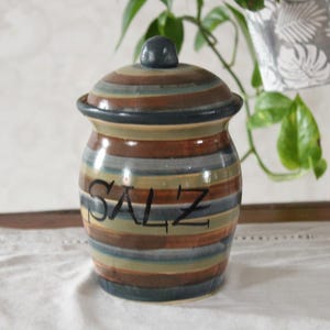 Könnte beinhalten: Ein Salzstreuer aus Keramik mit Deckel, mit horizontalen Streifen in Braun-, Blau- und Grüntönen. Das Wort "SALZ" ist in Schwarz auf die Vorderseite geschrieben. Das Gefäß steht auf einer weißen Oberfläche.