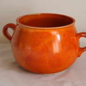 Könnte beinhalten: Ein kleiner orangefarbener Keramiktopf mit zwei Henkeln. Der Topf hat eine gesprenkelte Glasur und einen weißen Rand.