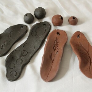 Op de afbeelding: Een verzameling handgemaakte klei-objecten, waaronder twee donkergrijze langwerpige stukken met cirkelvormige afdrukken, twee terracotta-kleurige langwerpige stukken met bladafdrukken en vier kleine bolvormige kralen in donkergrijs en terracotta.