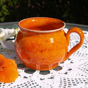 Może przedstawiać: Ręcznie wykonany, pomarańczowy ceramiczny kubek o zaokrąglonym korpusie i zakrzywionym uchwycie. Kubek ma błyszczące wykończenie i nakrapianą fakturę, z jaśniejszą krawędzią. Kubek stoi na białej koronkowej serwetce.