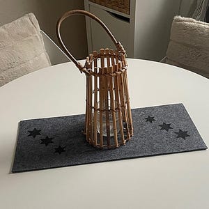 May include: A bamboo lantern with a handle stands on a grey felt placemat with black star cutouts. The lantern has a cylindrical shape and holds a glass candle holder. The placemat is on a white table.