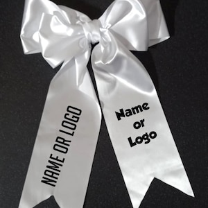 May include: A large, white satin bow with long, pointed streamers. The streamers display the text "NAME OR LOGO" and "Name or Logo" in black, block letters. The bow is set against a dark background.