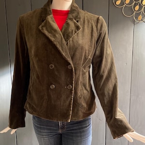 May include: A brown double-breasted blazer with four buttons. The blazer is made of a soft, velvety material and has a classic notched collar. The mannequin is also wearing a red top and blue jeans.