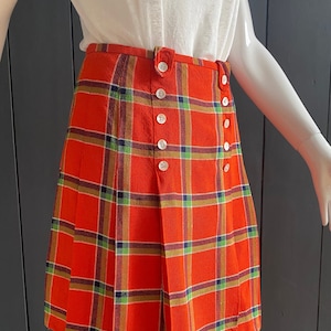 May include: A red tartan skirt with a button-up front and a pleated design. The skirt is made of woven fabric and has a short length.