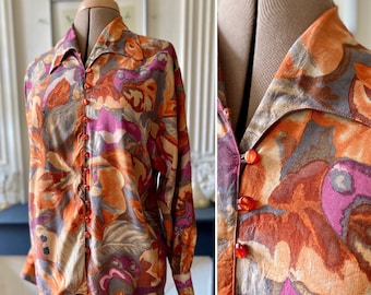 May include: A long-sleeved button-up shirt with a large abstract floral print in shades of orange, pink, and grey. The shirt has a wide collar and orange buttons.