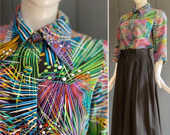 May include: A colourful, abstract print shirt with a black background and long sleeves. The shirt has a collar and button closure. The print features a variety of colours, including green, purple, orange, and yellow.