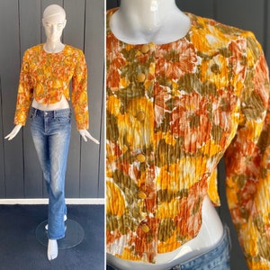 May include: A cropped, long-sleeve top with a floral pattern in shades of yellow, orange, and brown. The top has a round neckline and is buttoned down the front. The top is paired with blue jeans.