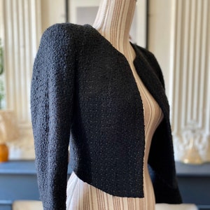 May include: A black cropped jacket made from textured tweed fabric. The jacket has a round neckline and long sleeves.