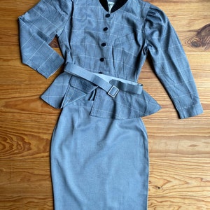 May include: A gray plaid skirt suit with a black velvet collar and a matching belt. The jacket has a fitted bodice and long sleeves. The skirt is a pencil skirt with a high waist.