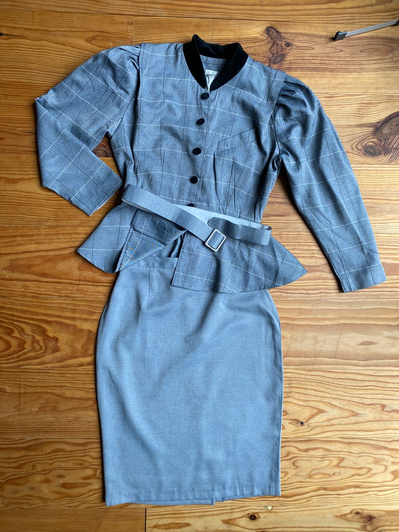 May include: A gray plaid skirt suit with a black velvet collar and a matching belt. The jacket has a fitted bodice and long sleeves. The skirt is a pencil skirt with a high waist.