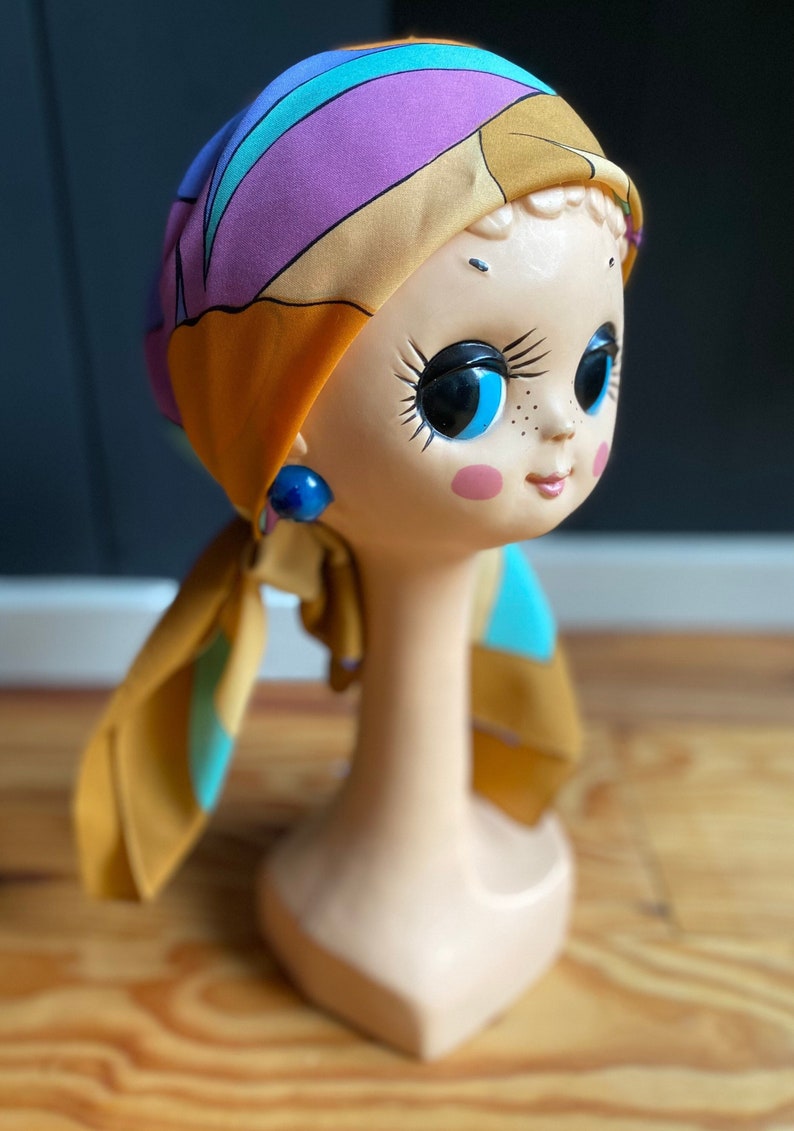 May include: A doll head with blue eyes and pink cheeks wearing a yellow, purple, and blue patterned headscarf. The doll is wearing blue earrings.