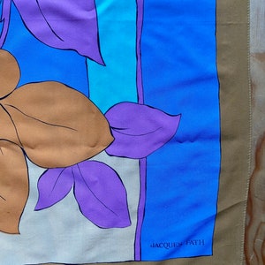 May include: A tan silk scarf with a floral design in shades of purple, blue, and orange. The scarf has a blue border and the text "JACQUES FATH" in white.