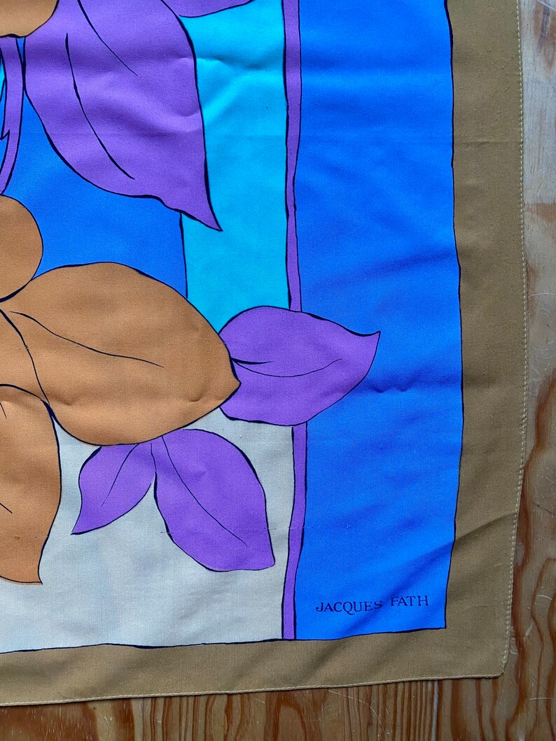 May include: A tan silk scarf with a floral design in shades of purple, blue, and orange. The scarf has a blue border and the text "JACQUES FATH" in white.