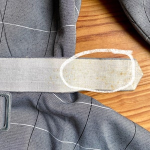May include: A close-up of a gray and white plaid fabric belt with a white fabric lining. The lining has a few brown stains.