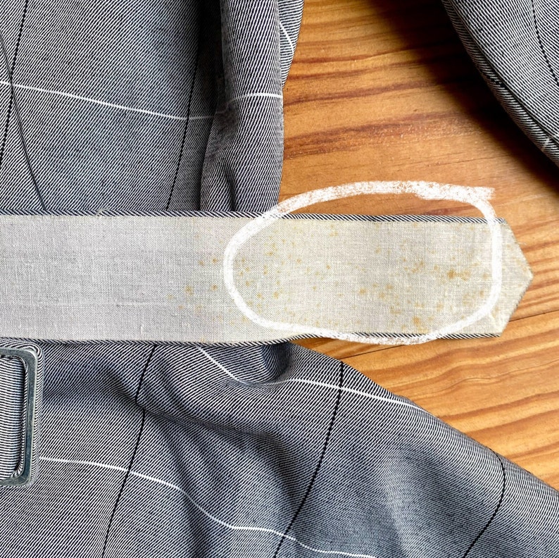 May include: A close-up of a gray and white plaid fabric belt with a white fabric lining. The lining has a few brown stains.