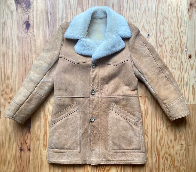 May include: A tan suede shearling coat with a large white shearling collar. The coat has a button closure and two large pockets.