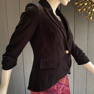 Peut inclure: Un blazer en velours noir avec un seul bouton doré et un revers cranté. Le blazer a des manches trois-quarts et une seule poche. La veste est présentée sur un mannequin.