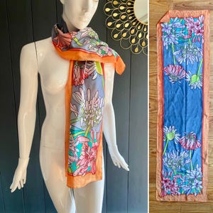 May include: A long, orange silk scarf with a floral print of pink, blue, and green flowers on a gray background. The scarf is tied around a mannequin's neck.