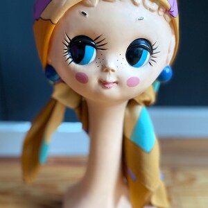 May include: A ceramic doll head with large blue eyes and a yellow, blue, and purple patterned scarf. The doll head is on a white stand.