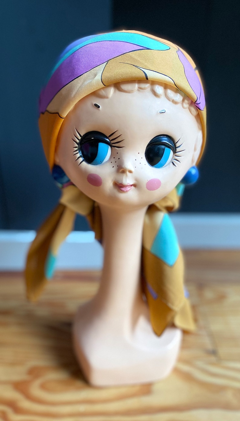 May include: A ceramic doll head with large blue eyes and a yellow, blue, and purple patterned scarf. The doll head is on a white stand.