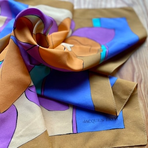 May include: A brown, blue, purple, and cream silk scarf with an abstract design. The scarf has the text "JACQUES FATH" printed on it.