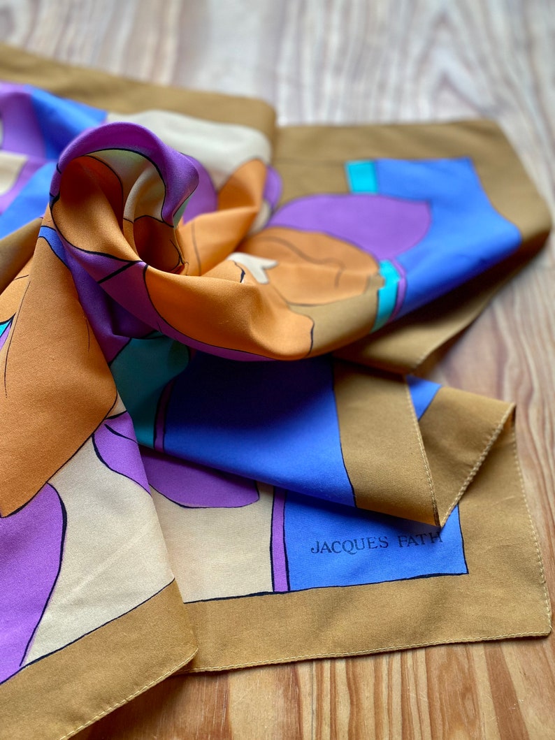 May include: A brown, blue, purple, and cream silk scarf with an abstract design. The scarf has the text "JACQUES FATH" printed on it.