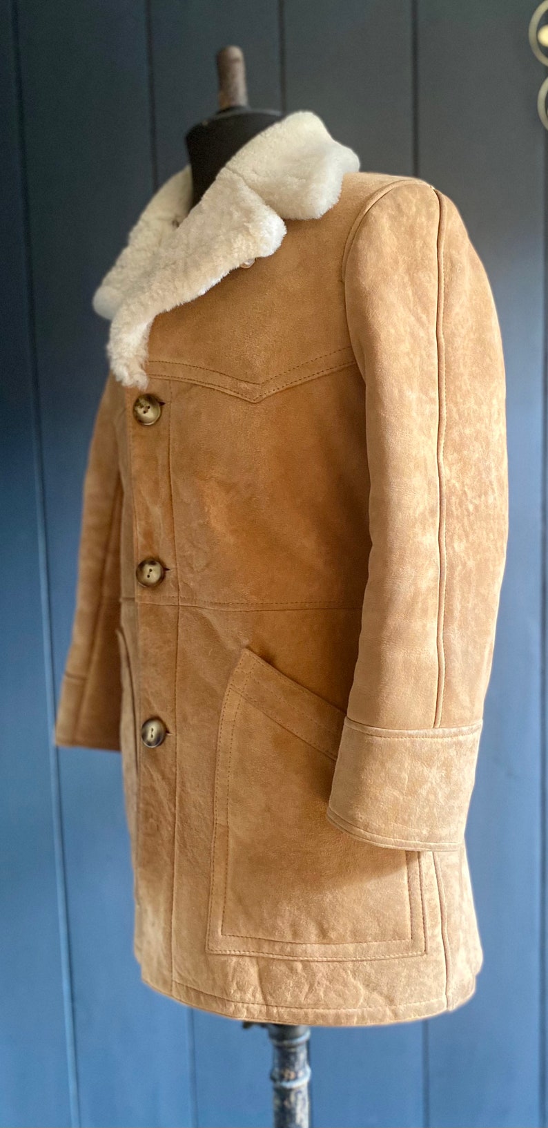 May include: A brown suede shearling coat with a white fur collar and a button closure. The coat has a patch pocket on the left side and a seam down the center of the back.