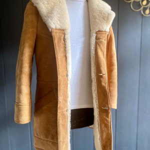 May include: A brown suede shearling coat with a white fur collar and lining. The coat has a button closure and two pockets.