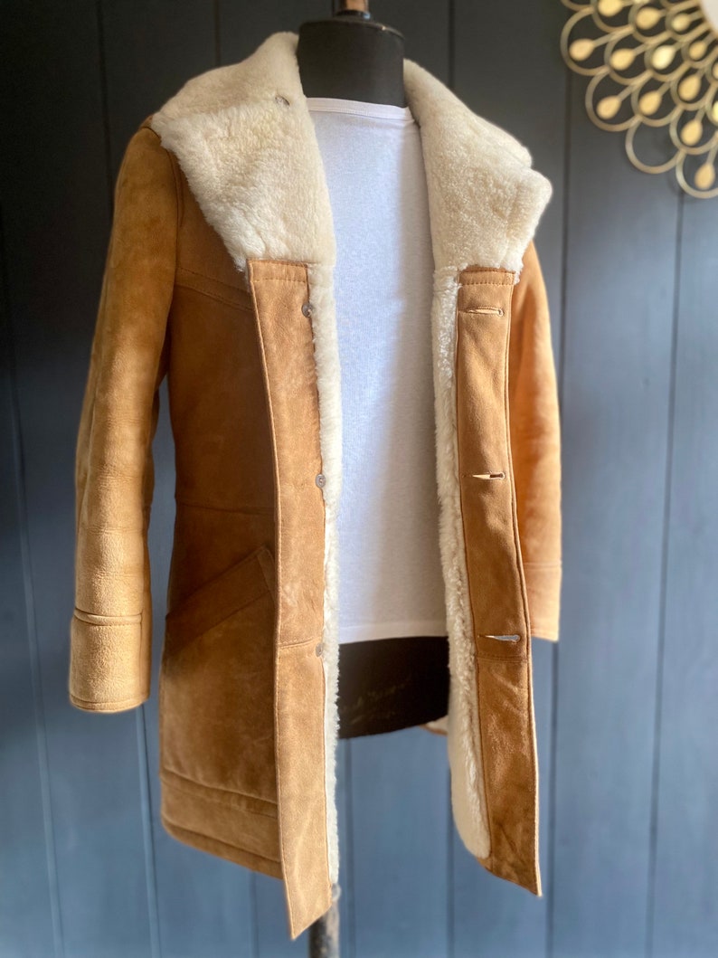 May include: A brown suede shearling coat with a white fur collar and lining. The coat has a button closure and two pockets.