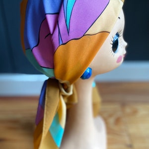 May include: A colorful patterned headscarf in shades of orange, purple, blue, and yellow. The scarf is tied in a knot at the back of the head and the ends are hanging down the back.