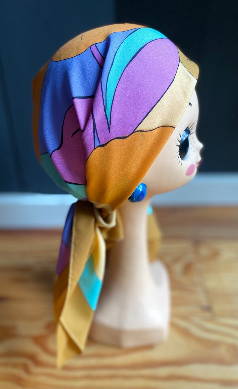 May include: A colorful patterned headscarf in shades of orange, purple, blue, and yellow. The scarf is tied in a knot at the back of the head and the ends are hanging down the back.