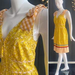 May include: Yellow sundress with a V-neck and a floral pattern. The dress features orange and white gingham straps and a matching hemline. The dress has a fitted bodice and a flared skirt, with a zipper closure.