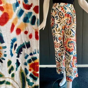 May include: Cream-colored pants with a vibrant, colorful floral pattern. The design features red, orange, blue, and green accents. The pants are displayed on a mannequin, showcasing the full length and fit of the garment.