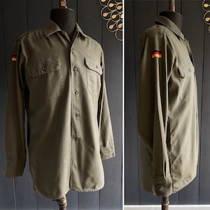 Peut inclure: Une chemise à manches longues de couleur verte avec un col boutonné et deux poches. La chemise a un patch du drapeau allemand sur la manche gauche.