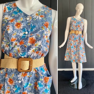 Peut inclure: Robe bleue sans manches ornée de fleurs blanches, orange et bleues. La robe a un col en V, des boutons blancs et une ceinture beige tressée avec une boucle carrée en bois. La robe est présentée sur un mannequin.