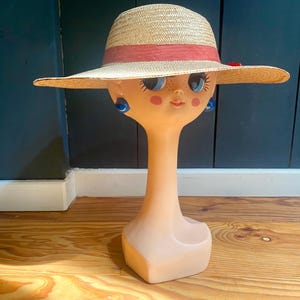 May include: A straw sun hat with a wide brim and a red band is displayed on a vintage mannequin head. The mannequin has blue eyes, rosy cheeks, and blue earrings. The hat is approximately 35 cm in diameter.