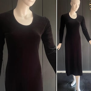 May include: A long, dark brown velvet dress with long sleeves and a scoop neckline. The dress is displayed on a mannequin, showcasing its form-fitting silhouette. The fabric has a subtle sheen, suggesting a luxurious feel. The dress is a classic, elegant design.