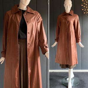 Può includere: Un trench coat marrone con collo e chiusura con bottoni. Il cappotto ha maniche lunghe con polsini abbottonati e orlo al ginocchio. Il cappotto è esposto su un manichino.