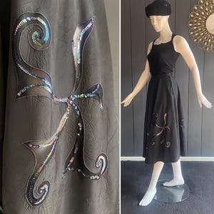 May include: A dark gray skirt with a large sequined design. The design is an abstract shape with iridescent sequins. A black top and hat are on a mannequin. The skirt has a flared shape.