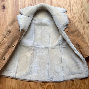 May include: A brown suede shearling coat with a white fur lining. The coat is open and the lining is visible. The coat has a button closure and two pockets.