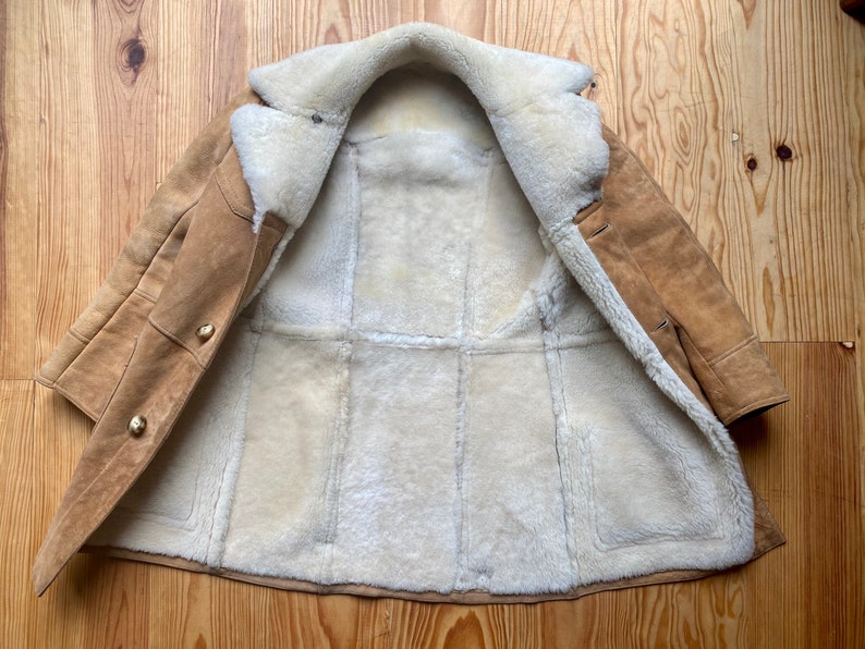 May include: A brown suede shearling coat with a white fur lining. The coat is open and the lining is visible. The coat has a button closure and two pockets.