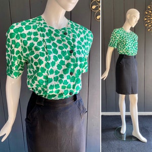 Puede incluir: Una blusa blanca con estampado abstracto verde y cierre de botones en la parte delantera. La blusa tiene mangas cortas y se combina con una falda negra.