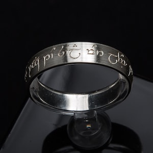Peut inclure: Une bague en argent avec une inscription dans une langue ancienne. L'inscription se lit comme suit : "Un anneau pour les gouverner tous, Un anneau pour les trouver, Un anneau pour les amener tous et dans les ténèbres les lier."