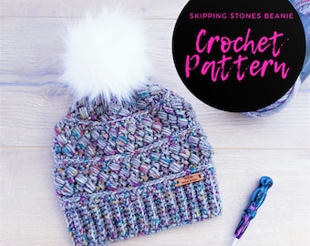 May include: A crochet beanie hat with a white pom pom, made with a multi-colored yarn. The beanie has a brown leather tag that says "Toffee Crochet Designs". The image also includes a crochet hook and a ball of yarn. The text "Skipping Stones Beanie Crochet Pattern" is visible in the image.