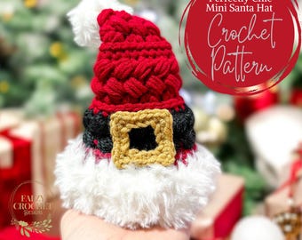 May include: A crochet pattern for a miniature Santa hat. The hat is red with a black and white belt and a gold buckle. The hat is sitting on a hand with a blurry background of a Christmas tree and presents.