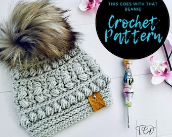 Puede incluir: Un gorro de ganchillo gris claro con un pompón de piel sintética. El gorro tiene un patrón de puntadas texturizadas y una pequeña etiqueta de cuero. La imagen incluye el texto "THIS GOES WITH THAT BEANIE Crochet Pattern". También se ve un ganchillo decorativo.
