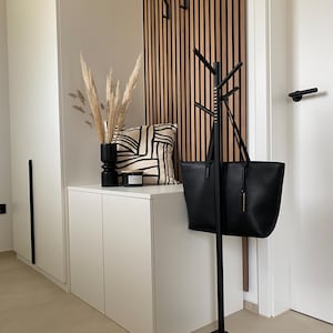 Op de afbeelding: Een moderne entree met een zwarte kapstok met een draagtas, een witte kast en een decoratief kussen. De muur heeft een houten lattenontwerp met zwarte haken. Een zwarte vaas bevat gedroogde planten.