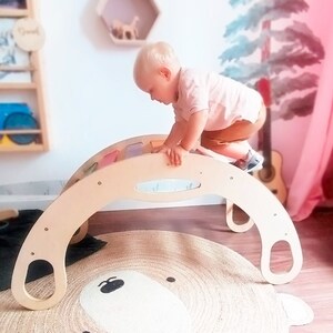 Peut inclure: Un enfant grimpe sur un bascule en bois avec des panneaux colorés. Le bascule est sur un tapis avec un motif d'ours.