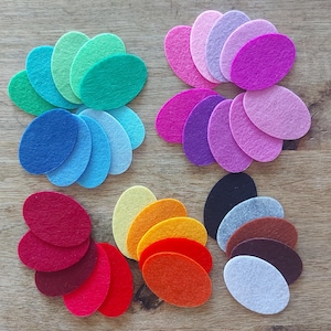 May include: Assortment of felt oval shapes in various colours, including red, orange, yellow, green, blue, purple, pink, brown, and grey. The felt shapes are arranged in a circular pattern on a wooden surface.
